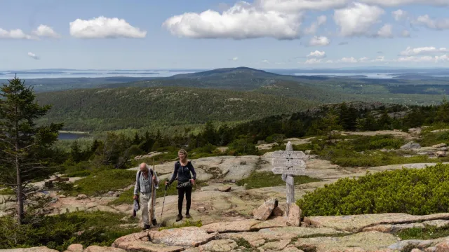 A man and woman holding walking sticks hike up a mountain summit near a wooden trail sign.