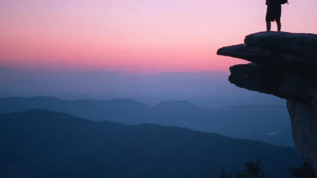 Silhouette of a man with backpack standing on McAfee Knob at sunset with mountains in the distance.