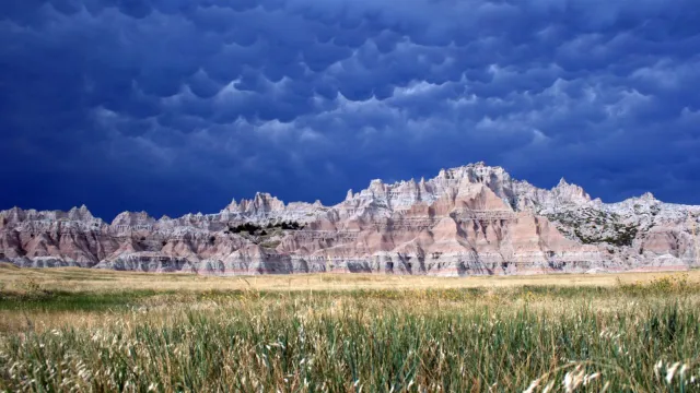 Layered badlands formations behind fields of green grass under cloudy and billowing clouds.