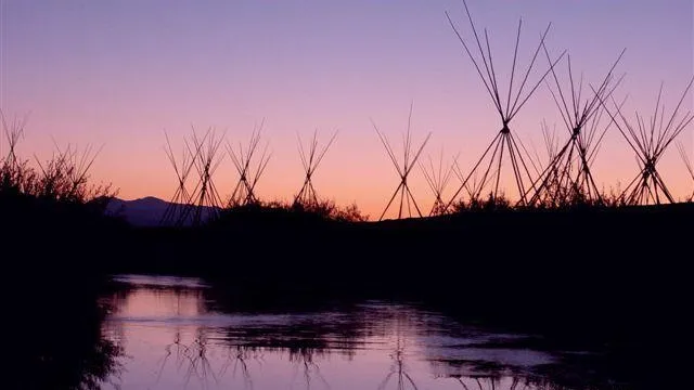Multiple tepee poles and a river are silhouetted against a dawn sky.