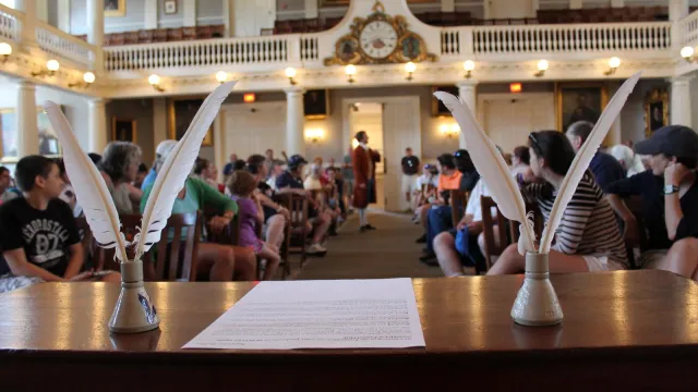 A desk with paper and two sets of quills frame a person in colonial wear standing in the aisle.