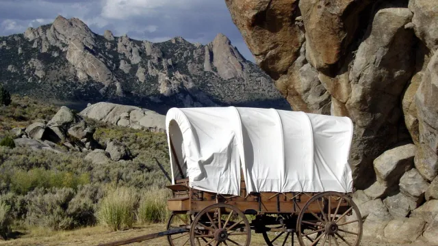 A white canvas wagon sits in front of a large rock buttress with mountains in the distance.