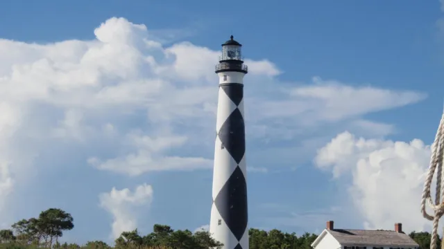 : Black & white patterned tower of the Cape Lookout Lighthouse stands against a blue & white cloud f