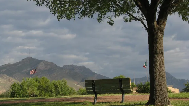 a bench next to a tree with U.S. and Mexican flags flying against a mountain background