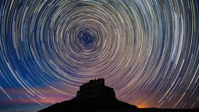 Fajada Butte's silhoutte shown under a timelapse image of stars appearing in a vibrant circle above.