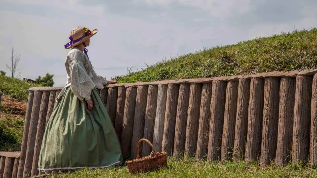 A woman in a 19th-century dress stands along an earthwork fortification
