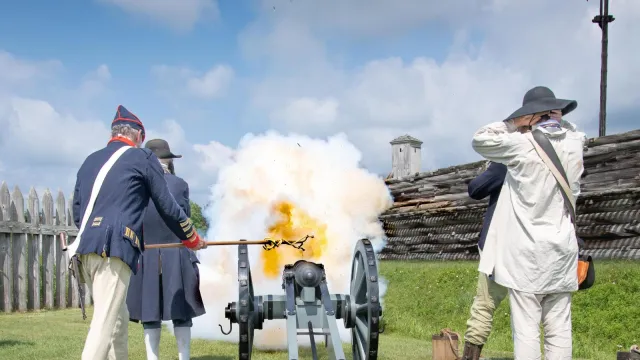 A cannon crew fires towards a wooden fort. An explosion of fire jets out in front of them.