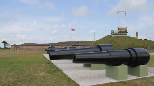 A row of cannon with Fort Moultrie in the background. A US flag is flying in the fort.