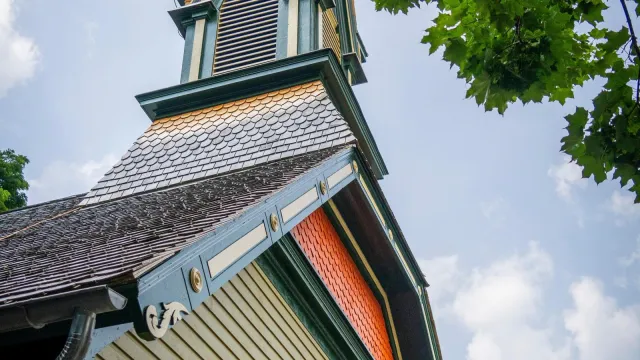 A tall, green and orange painted church steeple, against a blue sky, with a tree in the foreground.