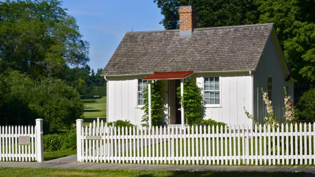 A one story wood frame house has board-and-batten exterior siding and a picket fence painted white.