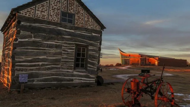 A historic cabin stands next to a concrete path that leads to a plow shaped visitor center
