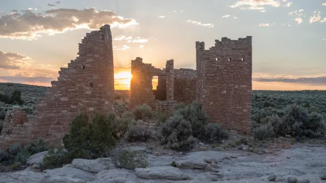 Tall stone structures with a setting sun shining through them. Desert landscape surrounds them.