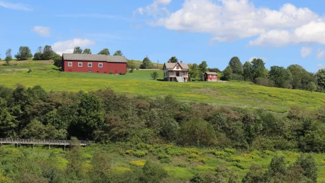 A farmhouse, shed, and long, red barn with windows atop a grassy hill
