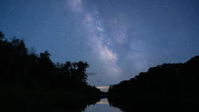 A blue night sky filled with stars sits over reflective water. Dark trees line the image.