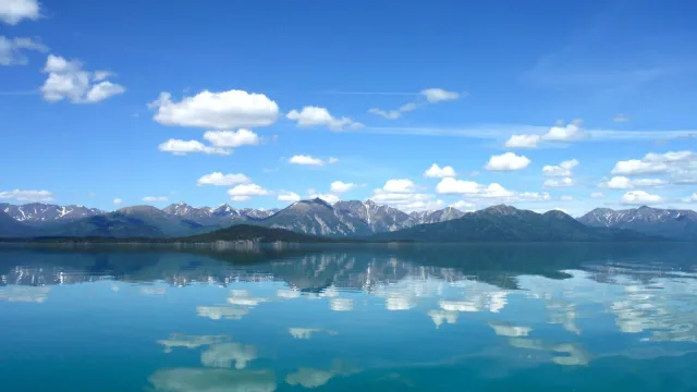 Photo of blue sky with fluffy white clouds reflect in calm lake with mountains in the background.