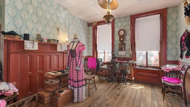 A room with wooden floors, blue wall paper, two windows, some furniture and a dress hanging.