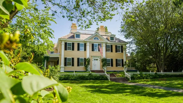 Yellow three-story mansion with symmetrical facade. Steps and large lawn in foreground. Framed by br
