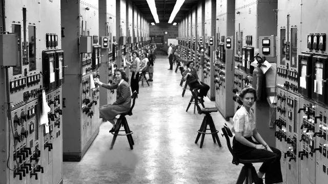 Several women sit on stools along a long hallway full of dials and meters.