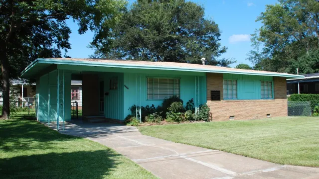 A small ranch-style home, aqua in color with light brown brick accents, sits on a shaded lot.
