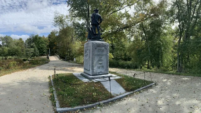The state of the Minute man stands atop a stone base. The wooden North Bridge is seen in the distanc