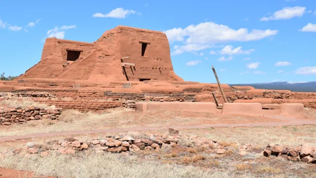 Large brown adobe mission church with low brick walls and kiva ladder in front