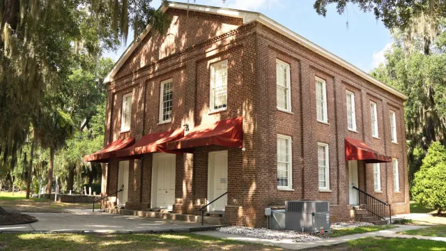 A two story brick building flanked by large live oak trees