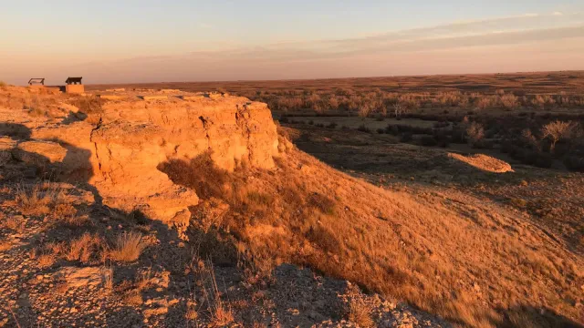 Warm light from the setting sun gives an orange glow to a rock bluff overlooking a grassland.