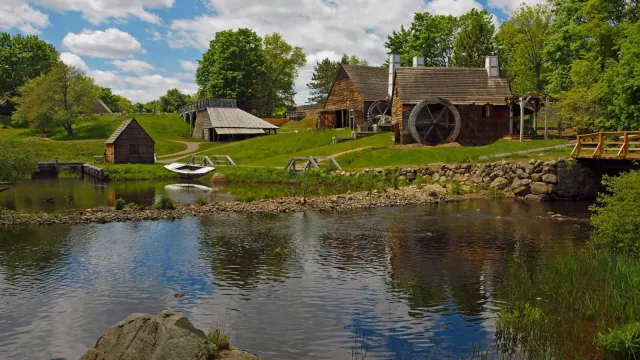 Several wooden structures amid green fields and trees under partly cloudy sky and beside river.