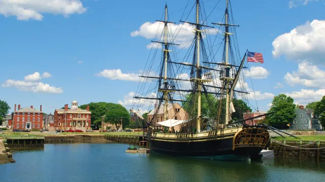 A three mast tall ship on the water under a blue sky with red brick buildings on the shore.