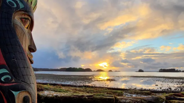 A totem pole in the foreground with a sunset over water in the background.