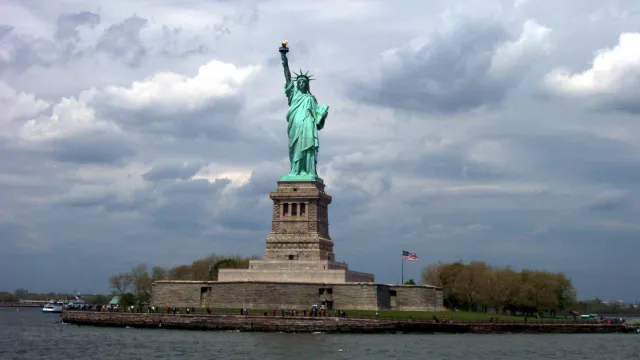 Patina green statue atop her pedestal on Liberty Island; grey cloudy sky in the distance
