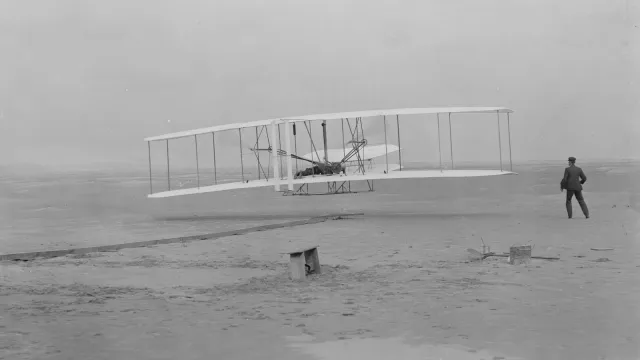 Orville takes to the air in the flyer for the first time as Wilbur assists.