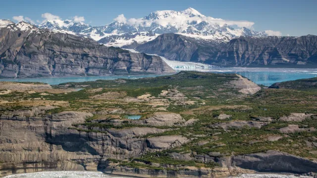 Glaciers loom over the ocean with large snowy mountains rising into blue skies